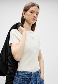 Λευκό ριγέ t-shirt με μικρή ετικέτα λογότυπου, συνδυασμένο με τζιν μπλε απόχρωσης που έχει υφή, κρατώντας μια μαύρη bomber jacket.