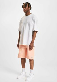 Tenue masculine comprenant un t-shirt en coton blanc ample et un short rose pastel, complété par des baskets blanches et des chaussettes blanches côtelées.