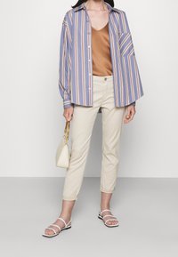 Camisa de botones a rayas en azul claro con rayas multicolores, camiseta color crema, pantalones crema, sandalias blancas y un pequeño bolso beige.