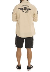Chemise beige à manches longues avec un design graphique noir représentant des ailes et un crâne, associée à un short cargo noir et des baskets noires.