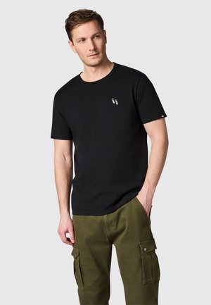 Man draagt een zwart T-shirt met korte mouwen met een klein kleurrijk slippers-ontwerp op de borst en olijfgroene cargobroek, staand met één hand in de zak.