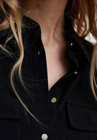 Chemise noire avec des accents de boutons dorés visibles, deux poches avant et un col. Le tissu semble lisse avec un léger éclat.