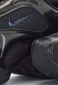 Gros plan d'une basket Nike noire montrant un mesh texturé, des panneaux synthétiques brillants et le logo Nike swoosh noir.