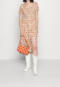 Bloemen midi-jurk met pofmouwen, licht materiaal met oranje en rode patronen. Gecombineerd met witte cowboylaarzen en een geruite oranje tas.