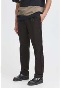 Pantalones negros de corte ajustado y textura rugosa, con parte frontal plana, bolsillos laterales y ajuste relajado. Combinados con zapatos negros brillantes.