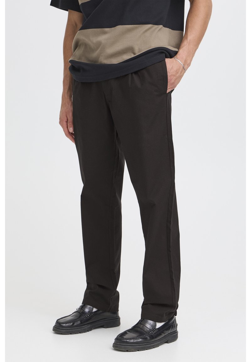 Pantalones negros de corte ajustado y textura rugosa, con parte frontal plana, bolsillos laterales y ajuste relajado. Combinados con zapatos negros brillantes.