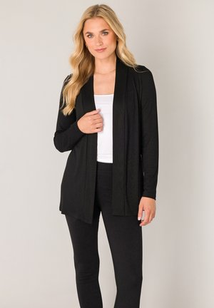 Blonde Frau, die eine schwarze offene Strickjacke, ein weißes Top und schwarze Hosen trägt, steht vor einem neutralen Hintergrund.