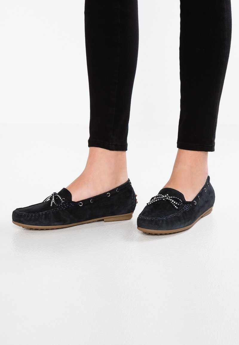 Mocassins en daim bleu marine avec nœud décoratif et accents métalliques, dotés d'une semelle plate en caoutchouc et de coutures le long des bords.