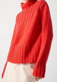 Person iført en lys rød chunky strikket turtleneck sweater med ribbede manchetter og kant, parret med lysebeige bukser.
