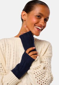 Femme souriante avec des cheveux plaqués en arrière, portant un pull crème en tricot torsadé et des gants en tricot sans doigts bleus marine, touchant son menton avec son doigt.