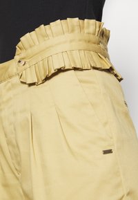 Pantalons en tissu jaune avec une taille froncée ornée de boutons et de plis. Agrémentés d'une petite étiquette en métal sur le côté.