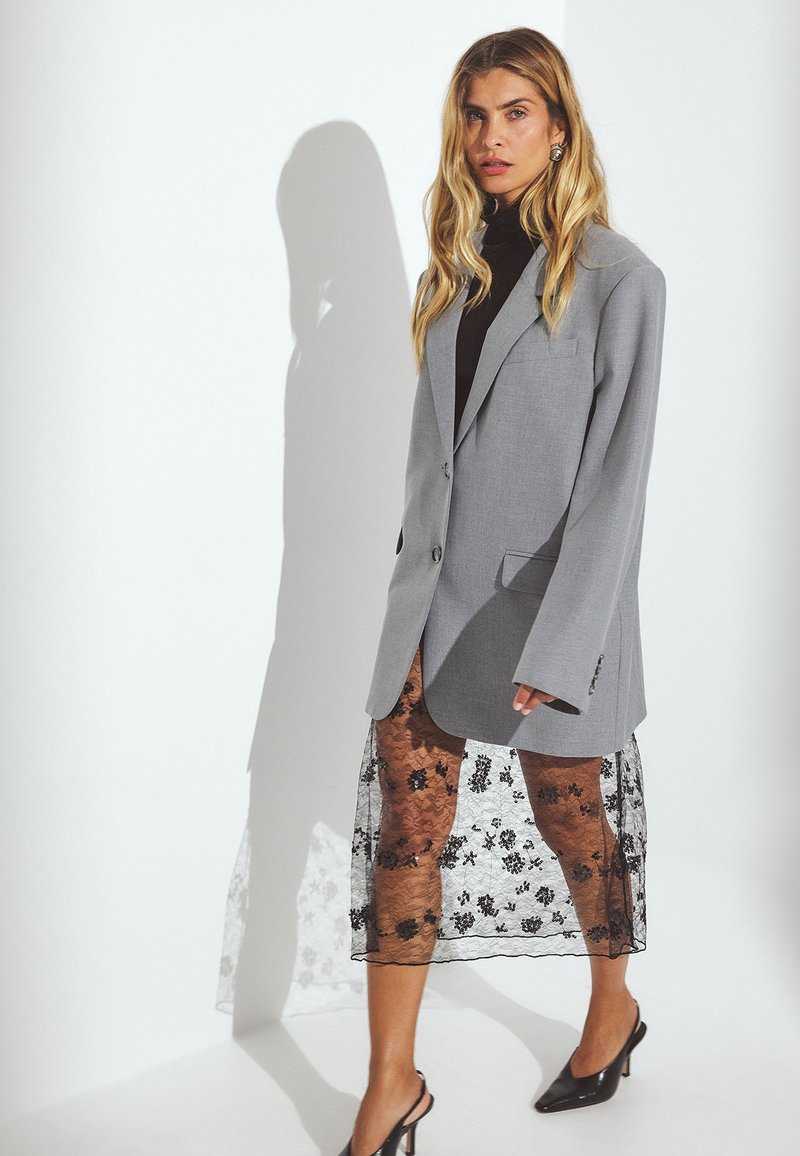 Blazer gris de gran tamaño con mangas largas, cierre de un solo botón, combinado con una falda de encaje negro transparente con patrones florales y mules de tacón alto.