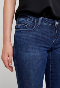 Pantalones vaqueros de mezclilla azul oscuro con una textura suave, que cuentan con un diseño ajustado, cinco bolsillos y un cierre de botón metálico.