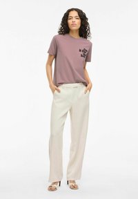 Camiseta morada clara con texto negro, pantalones crema de cintura alta y sandalias de tacón beige, sobre un fondo blanco liso.