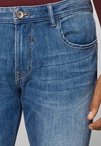 Lähikuva sinisistä farkuista, joissa on nähtävissä ompelu, metallinappi ja käsi taskun lähellä.