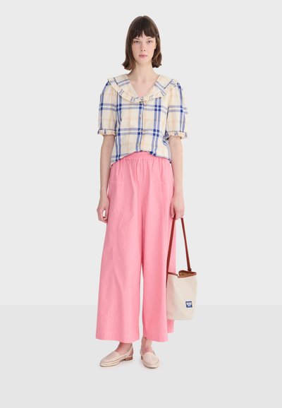 Femme portant un chemisier à carreaux bleu et beige avec un col à volants, un pantalon large rose, des mocassins beiges, tenant un sac fourre-tout beige avec des anses marron.