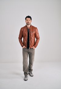 Brun læderjakke med lynlåslukning foran, stående krave og sidelommer, kombineret med en sort sweater, grå bukser og grå sneakers.