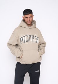 Béžová mikina s kapucňou a predným vreckom s veľkým textom "MULTPLY". Nosená s čiernymi teplákmi s malým logom. Hladký povrch.