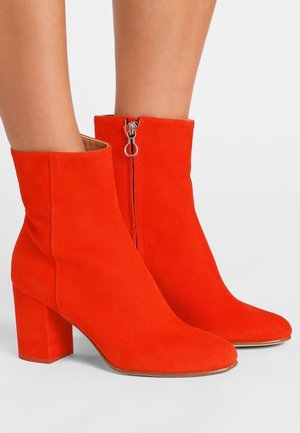 Czerwone zamszowe botki za kostkę na słupku z bocznym zamkiem, noszone na gołą stopę na jednolitym tle.
