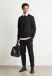 Sweatshirt preto de algodão com punhos canelados, combinado com calças de treino pretas com um logótipo. O modelo segura uma bolsa de couro preta e usa sapatos pretos.