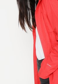 Veste légère rouge avec une finition texturée, dotée de manches longues, d'une capuche et d'une poche latérale. Le tissu est respirant et résistant à l'eau.