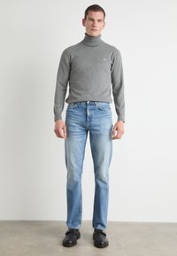 Pilkos spalvos turtleneck megztinis, prigludęs, minkštos tekstūros; šviesiai mėlynos tiesios kelnės; juodi loafersai su minimaliais akcentais.