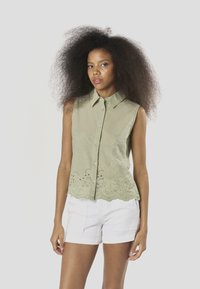 Heledad roheline varrukateta topp nööpidega, lainelise äärega ja alumisel osal tikitud lillemustriga, kombineeritud valgete lühikestega.