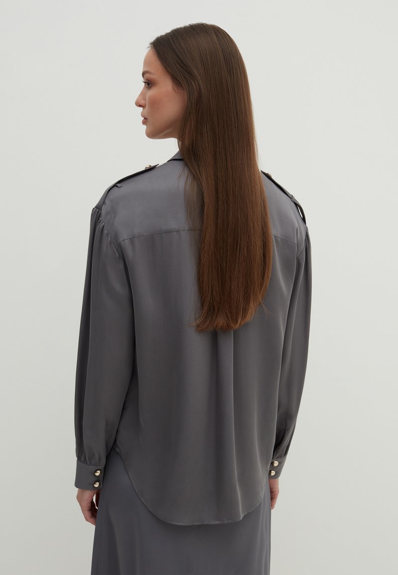 Blusa in satin grigia con colletto alto, spalline sulle spalle, maniche lunghe e bottoni decorativi sui polsini. Texture morbida e liscia.