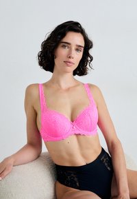 Rosa Spitzen-BH mit Blumenmuster, gepolsterten Cups und dekorativem Schleifchen. Das Model trägt hochgeschnittene schwarze Unterwäsche mit Spitzenakzenten.