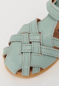 Lys turkis lær sandaler med vævet design, åben tå og lys brun sål. Inkluderer sømdetaljer langs kanterne.