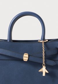 Blue suede handbag with smooth leather handles, gold hardware, a decorative charm, and a structured design featuring crossed detailing.
