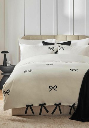 Beiges gepolstertes Bett mit weißer Bettwäsche mit schwarzen Schleifendesigns, schwarzen Kissen, schwarzem Überwurf, schwarzem Beistelltisch und einem Glas Wasser.