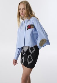 Camicetta azzurra chiara corta con taschino e motivo a quadretti. Mini gonna nera con motivi geometrici e dettagli hardware decorativi.