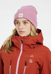 Rote wasserdichte Jacke mit hohem Kragen, pinken Reißverschlussdetails und schwarzem Zubehör. Pinker Strickmütze mit Logo-Patch auf der Vorderseite.