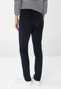 Persoon in een zwarte corduroy broek, witte sneakers en een zwart-wit gestreept shirt met lange mouwen, staand tegen een effen achtergrond.