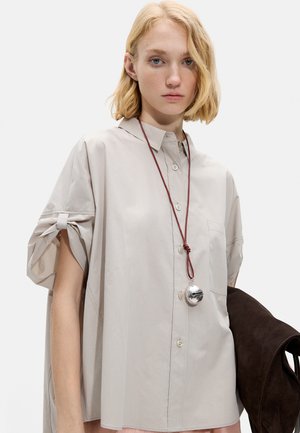 Młoda kobieta ubrana w luźną beżową koszulę zapinaną na guziki z zawiązanymi rękawami, srebrną zawieszką na czerwonym sznurku, trzymająca ciemnobrązową torbę zamszową.