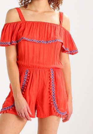 Mono rojo sin hombros con mangas acampanadas, que presenta patrones de diamante azules y acentos de pompones a lo largo del frente y los pantalones cortos.