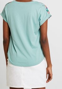 T-shirt vert clair à manches courtes avec des ourlets roulés, ornée d'une broderie florale sur les épaules. Associé à une jupe en denim blanche.