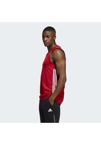 Débardeur de sport rouge avec des rayures blanches sur les côtés, associé à un pantalon noir arborant un logo Adidas blanc et des lignes d'accent.