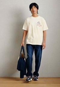 Κρέμ βαμβακερό μπλουζάκι με γραφική εκτύπωση, σκούρο μπλε τζιν και πολυχρωμα sneakers. Κουβαλώντας μια ναυτική τσάντα ώμου με ανοιχτόχρωμο εσωτερικό.