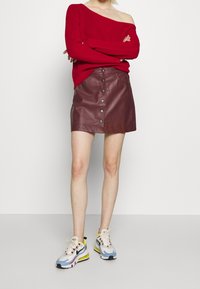 Czerwona prążkowana sweterka z odkrytymi ramionami nad bordową mini spódniczką skórzaną z przodu zapinana na zatrzaski, w połączeniu z kolorowymi sneakersami.
