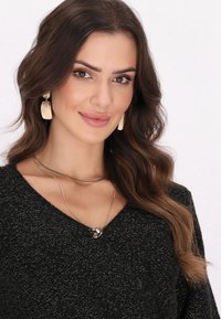 Maglione nero a trama con scollo a V, collane in argento sovrapposte e orecchini con nappine dorate. Capelli castani con onde morbide.