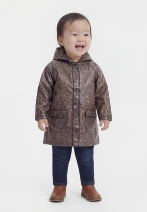 Copil mic zâmbind, purtând un palton cu glugă, imprimeu de leopard maro, blugi închiși la culoare și cizme maro, stând pe un fundal alb simplu.