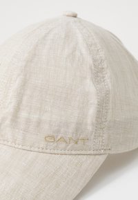Beige baseball sapka, texturált anyagból készült, finom varrással és arany hímzett logóval. Hajlított karimával tervezve.