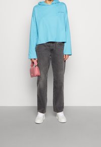 Hoodie court bleu clair avec un logo sur le devant, associé à un jean droit gris foncé. Baskets blanches et un petit sac à main rose avec une bandoulière.