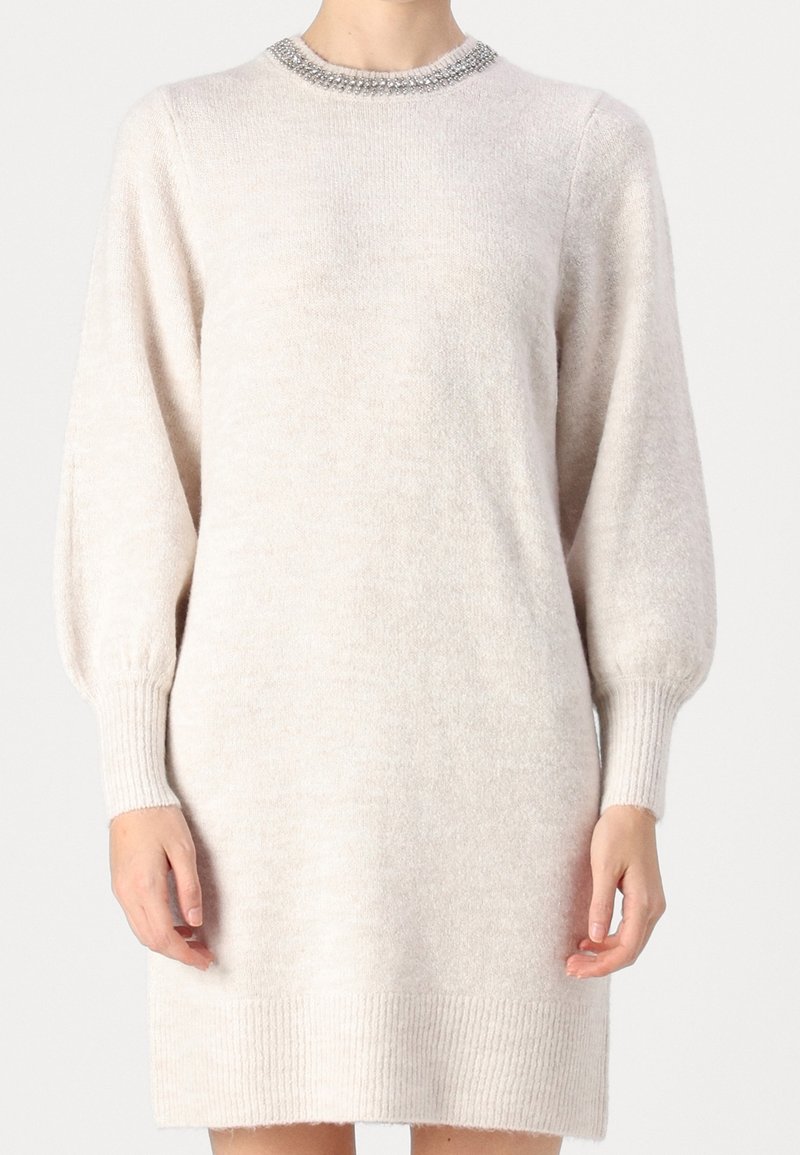 Femme portant une robe en maille beige clair à manches longues avec poignets et ourlet côtelés, et un col décoratif orné de perles.