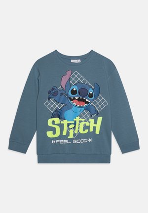 Blå langærmet sweatshirt med en grafik af Stitch og teksten "STITCH FEEL GOOD" i lysende grøn og hvid på en gitterbaggrund.