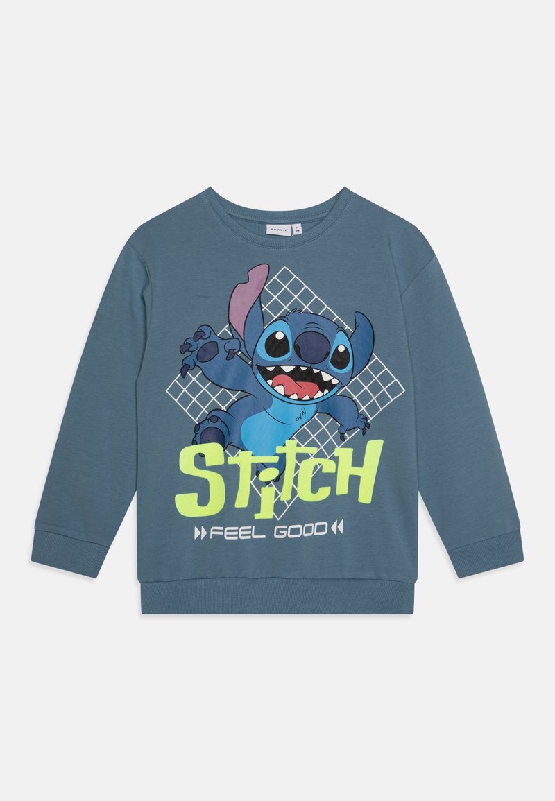 Sweat-shirt bleu à manches longues avec un graphisme de Stitch, accompagné du texte "STITCH FEEL GOOD" en vert vif et blanc sur un fond à carreaux.