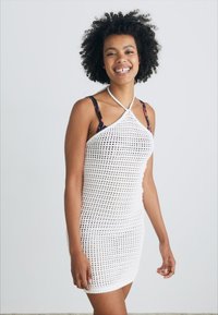 Bílá háčkovaná šaty s přiléhavým střihem, designem za krk a průsvitnou texturou. Obsahuje nastavitelné ramínka.