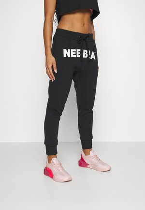 Kvinde iført sorte joggers med "NEBBIA"-logo, kort sort top og pink sportssko, stående på en grå gulv.